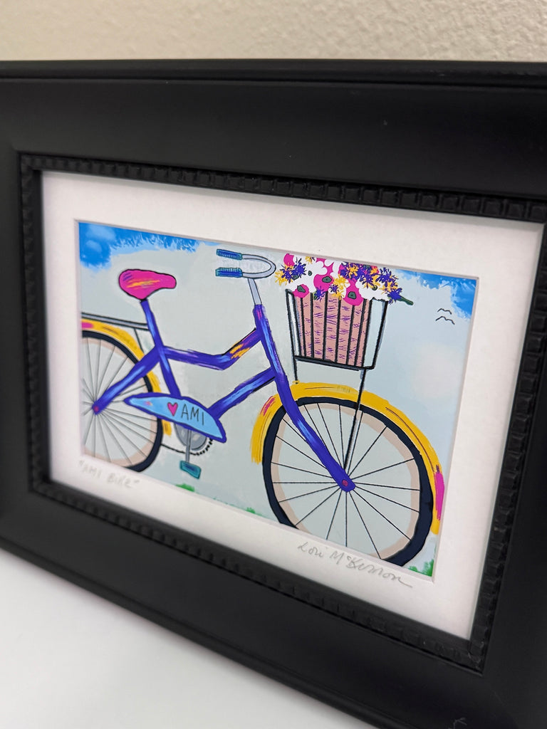 A framed art print featuring a colorful illustration of a purple bicycle with a flower basket, set in a black frame with a white mat.