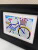 A framed art print featuring a colorful illustration of a purple bicycle with a flower basket, set in a black frame with a white mat.