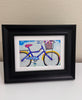 A framed art print featuring a colorful hand-painted style bicycle with a basket of flowers, displayed in a black frame on a white surface against a beige wall.