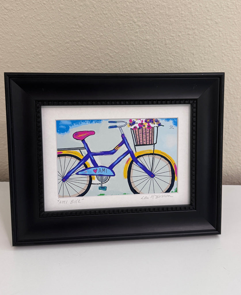 A framed art print featuring a colorful hand-painted style bicycle with a basket of flowers, displayed in a black frame on a white surface against a beige wall.