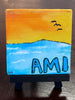 Small painted canvas with a sunset over water scene and 'AMI' text on a wooden stand.