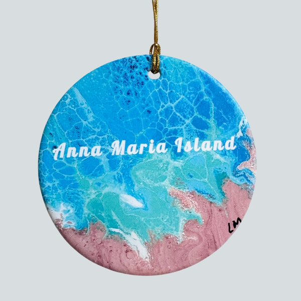 Round ceramic ornament featuring an abstract ocean design with blue water, turquoise waves, sand, and the text 'Anna Maria Island' in white script. The ornament hangs from a gold string against a light gray background.