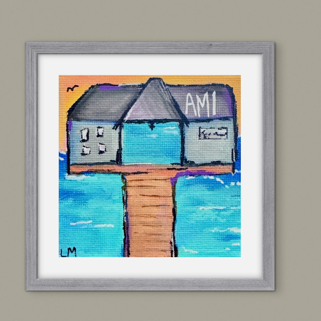 Framed artwork of a building on a pier with 'AMI' branding, set against a beach scene.