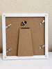 The back of a square white picture frame with a brown particle board backing, four silver metal hanging clips, and a central cardboard insert, shown against a beige wall.