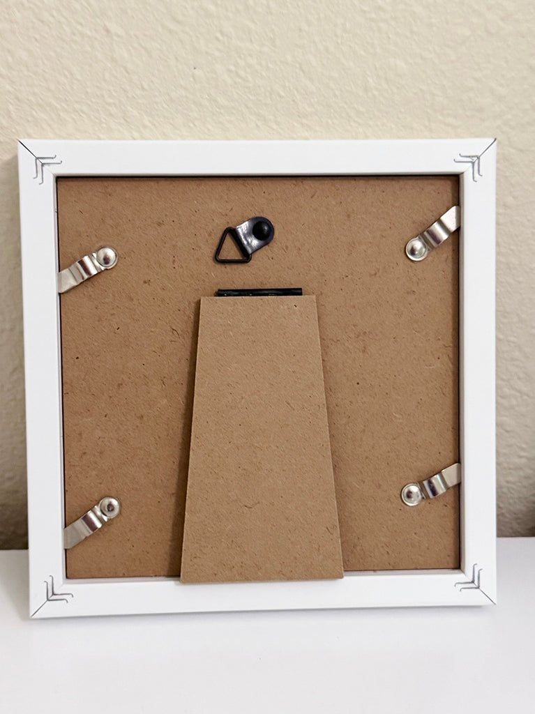 The back of a square white picture frame with a brown particle board backing, four silver metal hanging clips, and a central cardboard insert, shown against a beige wall.