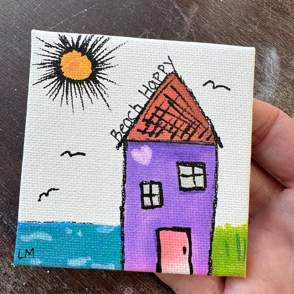 A small hand-painted canvas print of a whimsical house with the words 'Beach Happy' and a sun, held in a person's hand over a dark wooden surface.