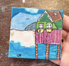 A small square canvas painting of a colorful beach house on stilts, held by a hand over a wooden surface. The painting features a green house with a pink porch, blue sky and water, and a sandy beach.