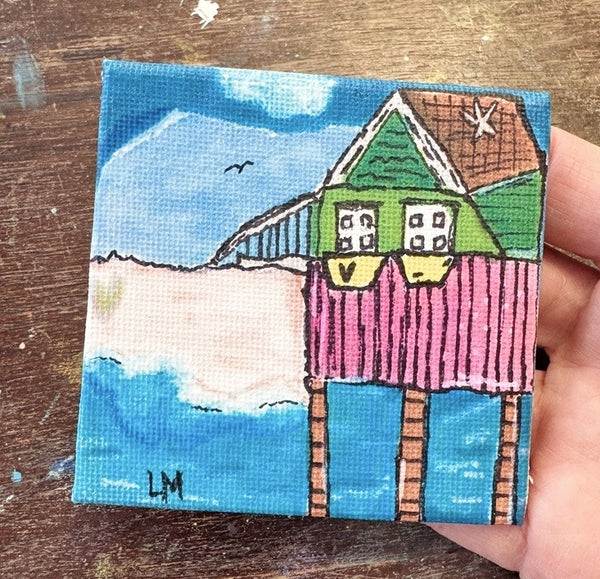 A small square canvas painting of a colorful beach house on stilts, held by a hand over a wooden surface. The painting features a green house with a pink porch, blue sky and water, and a sandy beach.