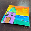 A small square canvas painting of a colorful house on a dark wooden table. The painting features a purple house with a green roof, a blue ocean, a yellow and orange sky, and green foreground, signed 'LM'.