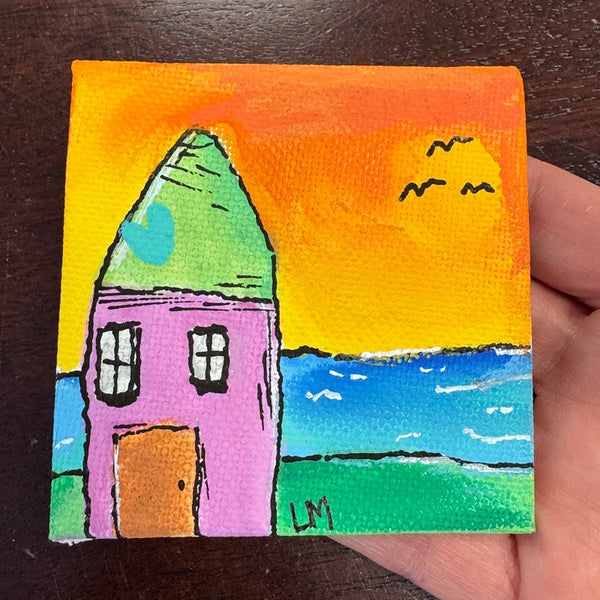A small square canvas painting of a colorful house at sunset, held in a hand over a dark wooden surface. The painting features a pink house with a green roof, a bright orange sky, blue water, and green land.