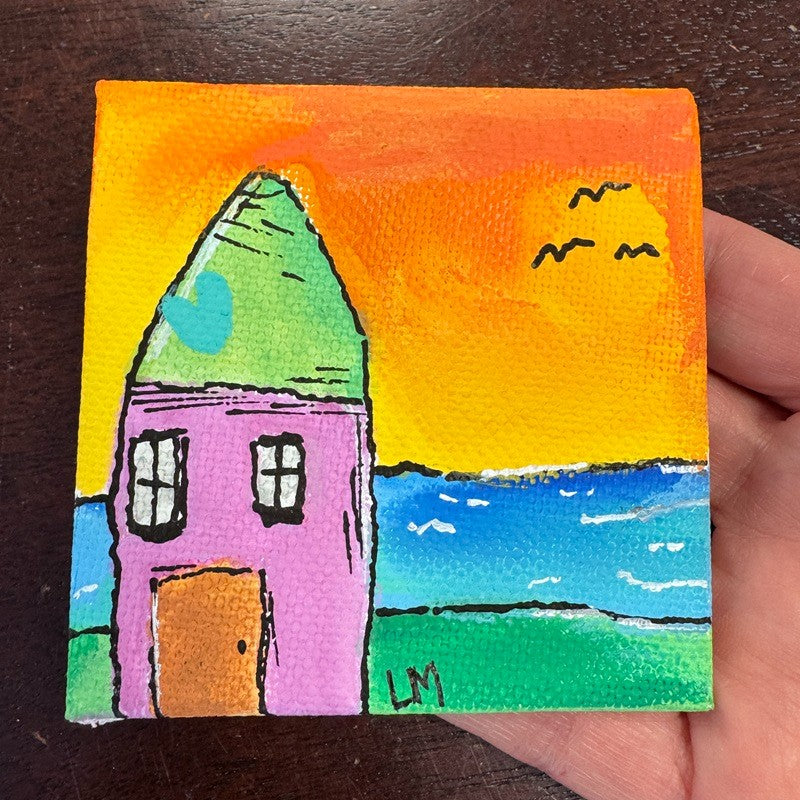 A small square canvas painting of a colorful house at sunset, held in a hand over a dark wooden surface. The painting features a pink house with a green roof, a bright orange sky, blue water, and green land.