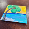 A square hand-painted canvas depicting a tropical beach scene with a palm tree, yellow sky, blue ocean, and white shoreline, resting on a dark wooden surface.