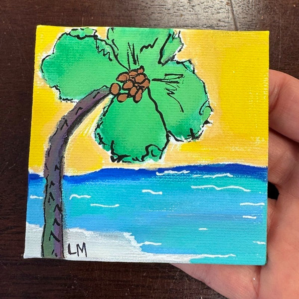 A hand holding a small square canvas painting of a tropical beach scene with a green palm tree, yellow sky, blue ocean, and white surf. The painting has a visible canvas texture and is signed 'LM' in the lower left corner.