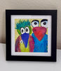 A framed colorful print of two whimsical birds displayed on a white surface against a textured beige wall. The left character has green spiky hair, a purple beak, and a pink/magenta body.