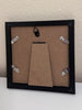 The back of a square black picture frame with a brown cardboard insert and four silver metal clips, shown against a textured off-white wall.