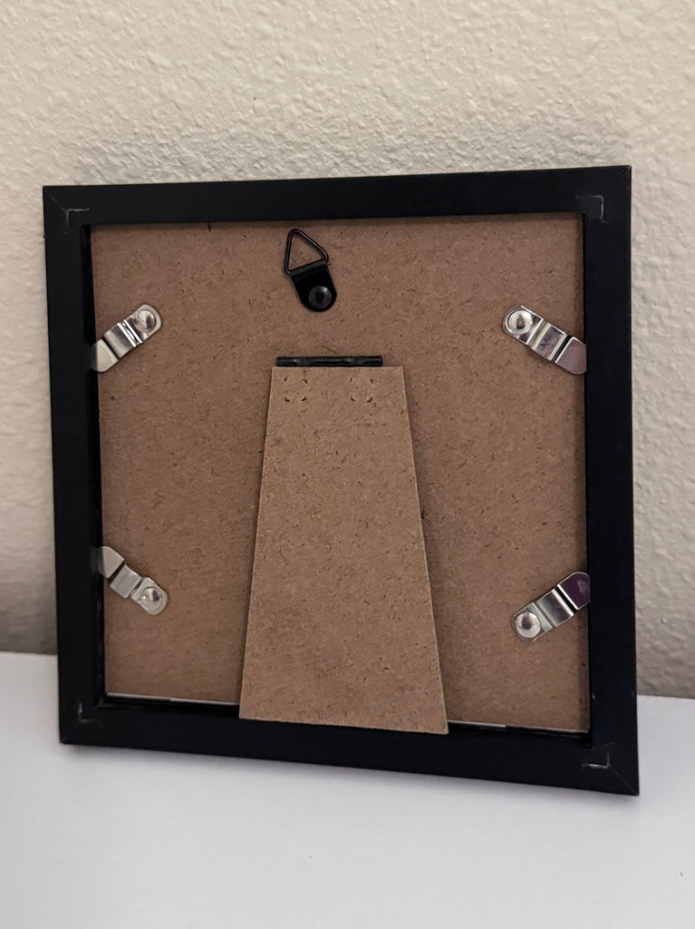 The back of a square black picture frame with a brown cardboard insert and four silver metal clips, shown against a textured off-white wall.