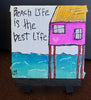 A small hand-painted canvas art piece on a black easel stand, featuring a whimsical pink house on stilts with a yellow roof, turquoise water, and the handwritten text 'Beach Life is the best Life' on a white background.