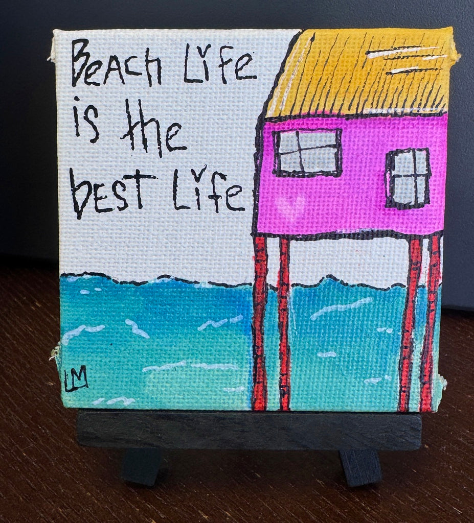 A small hand-painted canvas art piece on a black easel stand, featuring a whimsical pink house on stilts with a yellow roof, turquoise water, and the handwritten text 'Beach Life is the best Life' on a white background.