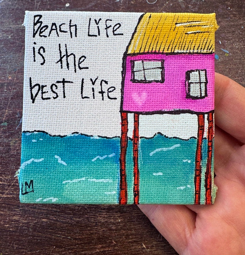 A square canvas painting held in a hand, featuring a pink house on stilts over blue water with a yellow roof, plus the text 'Beach Life is the best Life' on a white background.