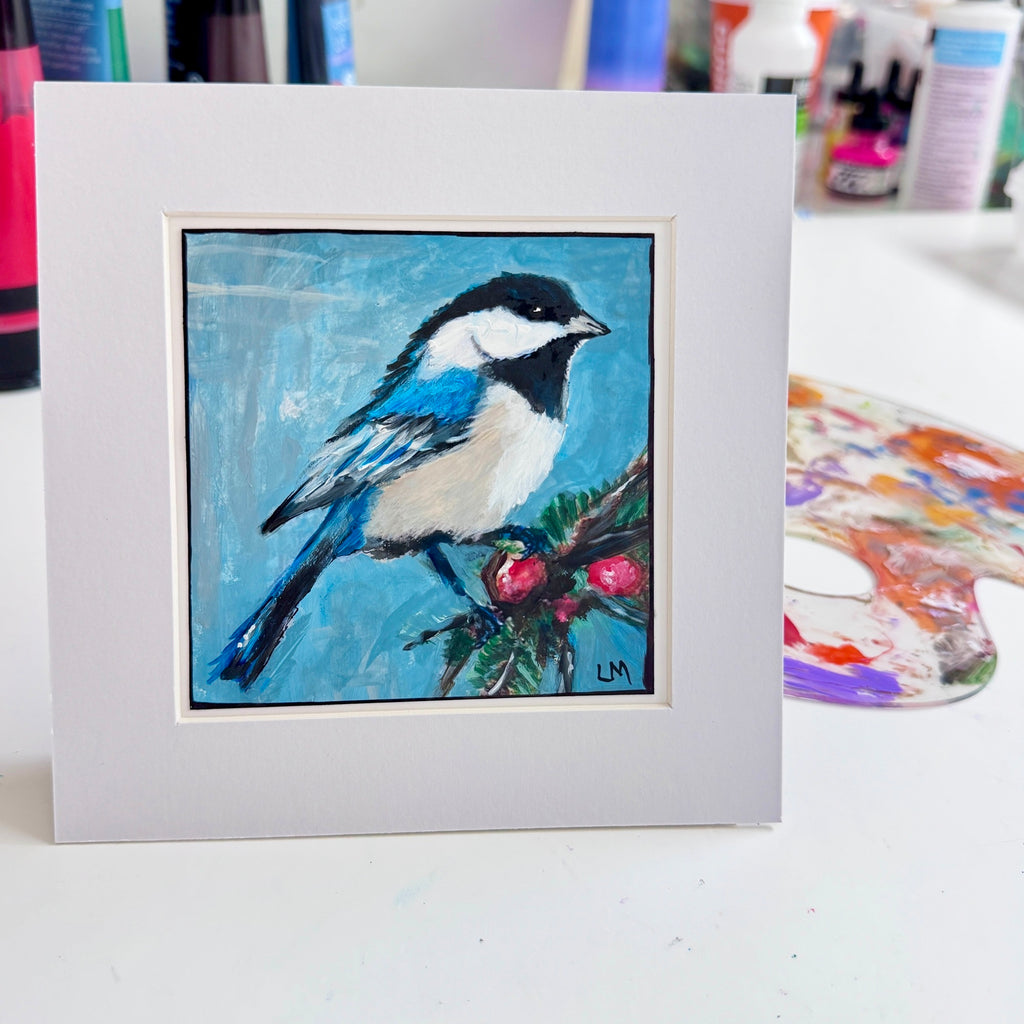 An acrylic bird painting in a white mat, displayed on an art studio table beside a paint palette