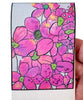 A close-up of a hand holding a magnetic bookmark featuring a floral pattern in pink, magenta, purple, and green with black outlines on a white paper background.