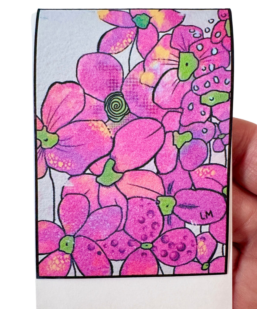 A close-up of a hand holding a magnetic bookmark featuring a floral pattern in pink, magenta, purple, and green with black outlines on a white paper background.