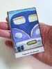 A hand holding a small rectangular magnetic bookmark featuring a stylized blue and purple van illustration with the word "BEACH" on a white card labeled "Magnetic Bookmark.".