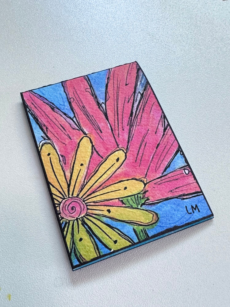 An art print featuring a colorful hand-drawn flower design, displayed on a light gray textured surface. The artwork depicts a yellow flower with a pink center and large pink petals on a blue background, signed 'LM' in the bottom right corner.