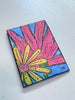 An art print featuring a colorful hand-drawn flower design, displayed on a light gray textured surface. The artwork depicts a yellow flower with a pink center and large pink petals on a blue background, signed 'LM' in the bottom right corner.