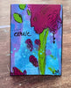 A small painting resting on a wooden surface, featuring an abstract floral design with a large magenta flower, green stem and leaves, blue and purple background washes, and the handwritten word "exhale" on the left.
