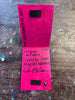 An open handmade artwork on a paint-splattered wooden table. The pages are a vibrant magenta-pink color with black handwritten text, signatures, and small black rectangular inserts, set against a dark brown wood background.