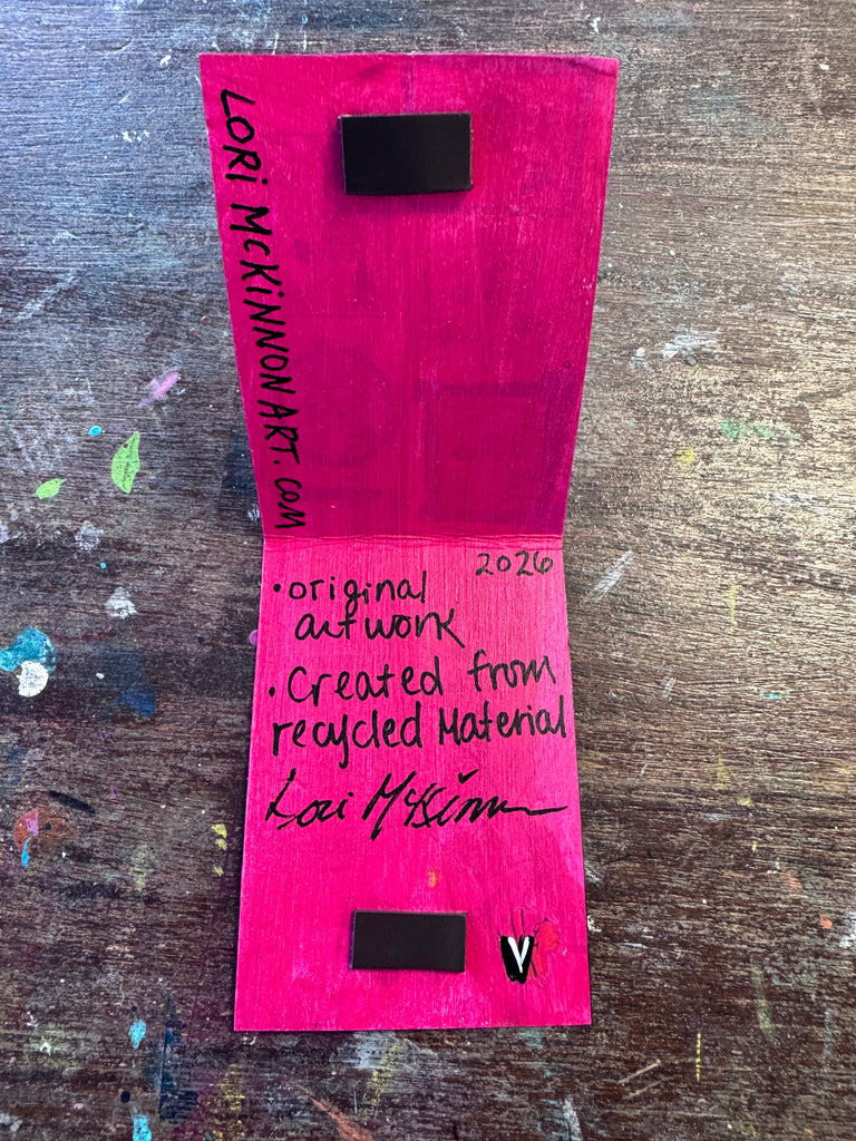 An open handmade artwork on a paint-splattered wooden table. The pages are a vibrant magenta-pink color with black handwritten text, signatures, and small black rectangular inserts, set against a dark brown wood background.
