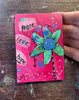 A hand holding a small square pink artwork card featuring a green flower with a blue face, hand-drawn text reading peace, love, and hope, on a worn wooden tabletop.