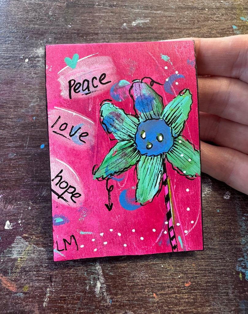 A hand holding a small square pink artwork card featuring a green flower with a blue face, hand-drawn text reading peace, love, and hope, on a worn wooden tabletop.
