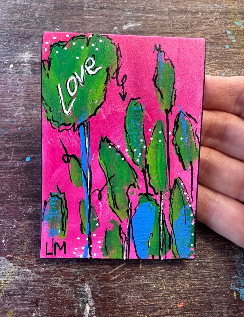 A small colorful painting on a wooden surface, held by a hand. The artwork features green leaf-like shapes with black outlines on a bright pink background, with the word 'Love' written in white inside one leaf.