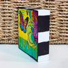 colorful butterfly painting on a wood block showing the sides are painted black and white. It is pictured on a table with a basket behind it