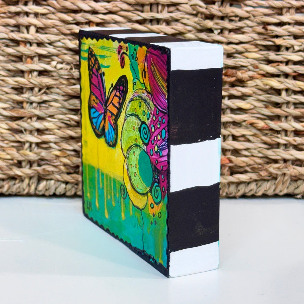 colorful butterfly painting on a wood block showing the sides are painted black and white. It is pictured on a table with a basket behind it