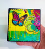 butterfly painting on a wood block with vivid colors of yellow, green, pink and blue. The artwork is held by a hand