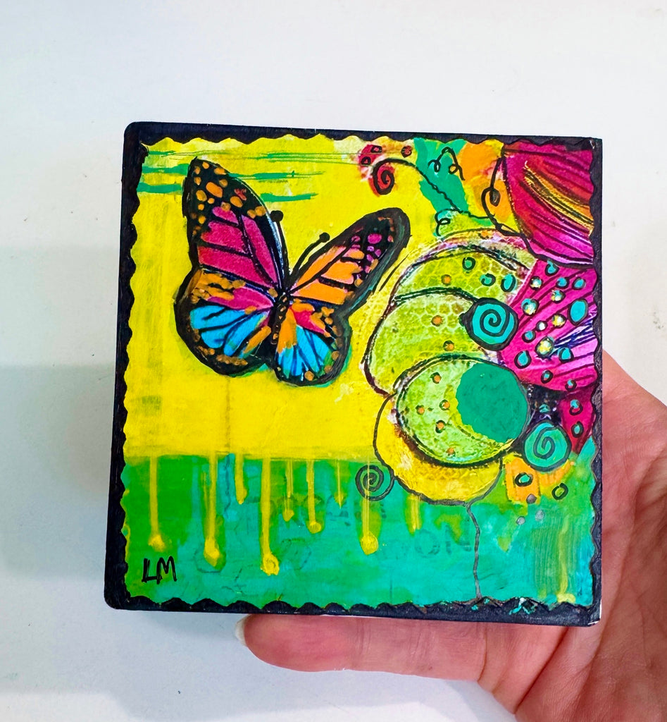 butterfly painting on a wood block with vivid colors of yellow, green, pink and blue. The artwork is held by a hand