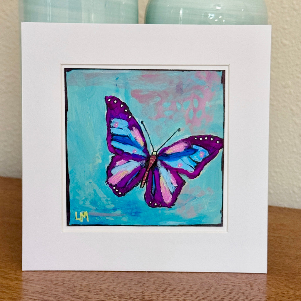 A butterfly painting in a white mat standing on a table with vases behind it
