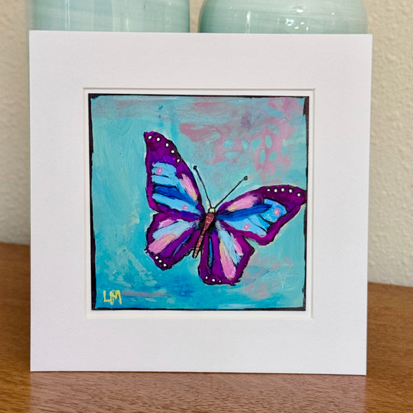 A butterfly painting in a white mat standing on a table with vases behind it