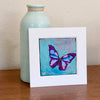 A butterfly painting in a white mat standing on a table with a vase behind it