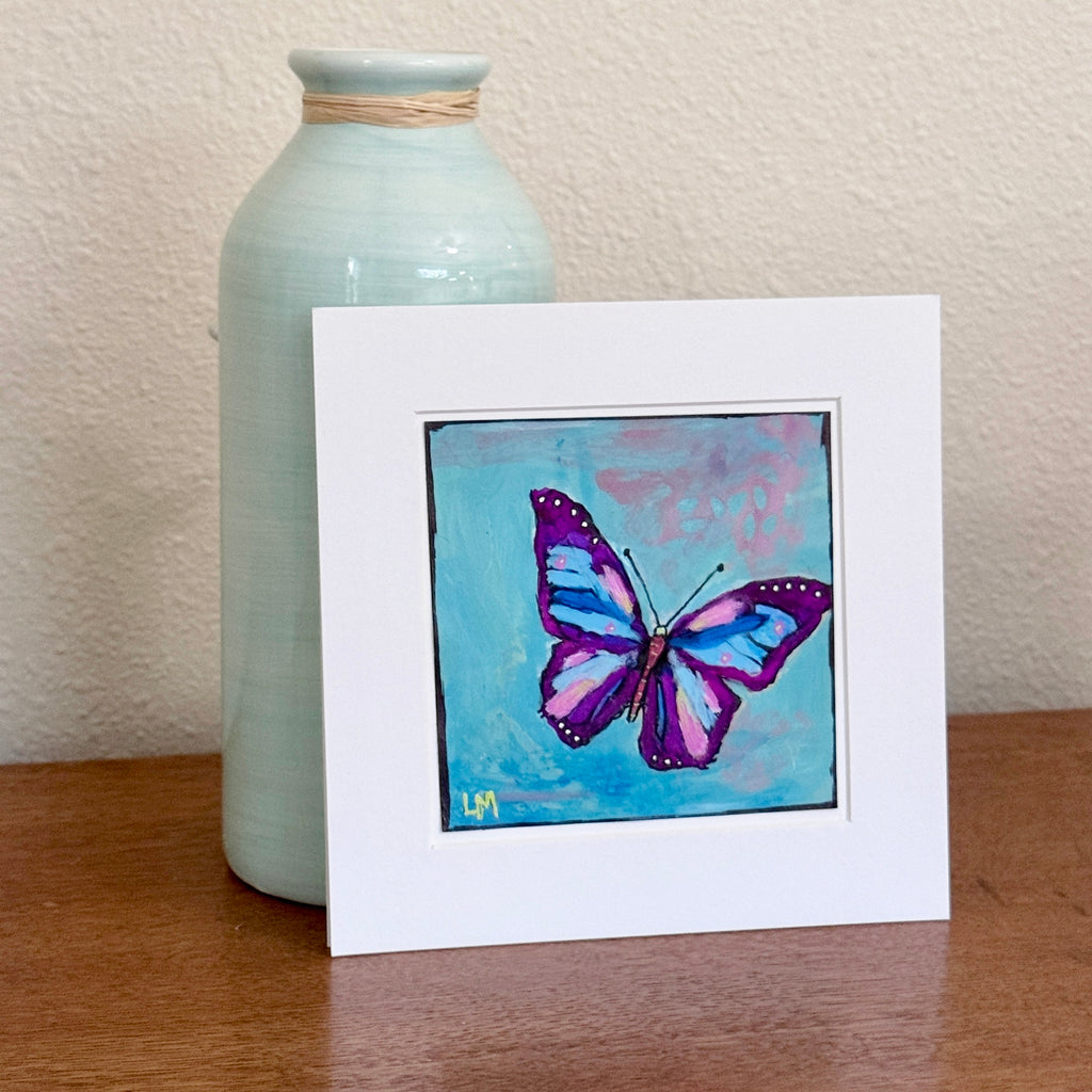 A butterfly painting in a white mat standing on a table with a vase behind it