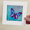 A butterfly painting in a white mat being held in a hand against a light beige wall