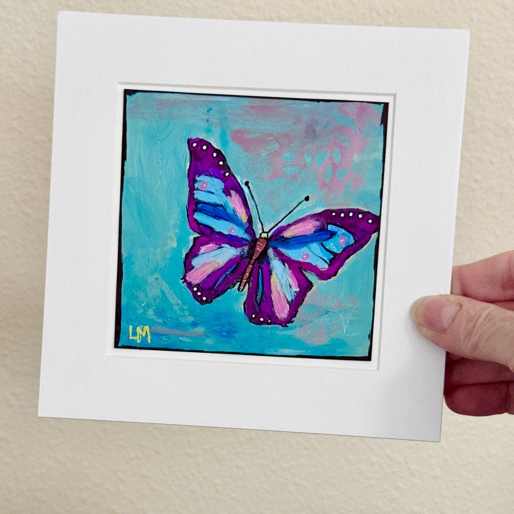 A butterfly painting in a white mat being held in a hand against a light beige wall