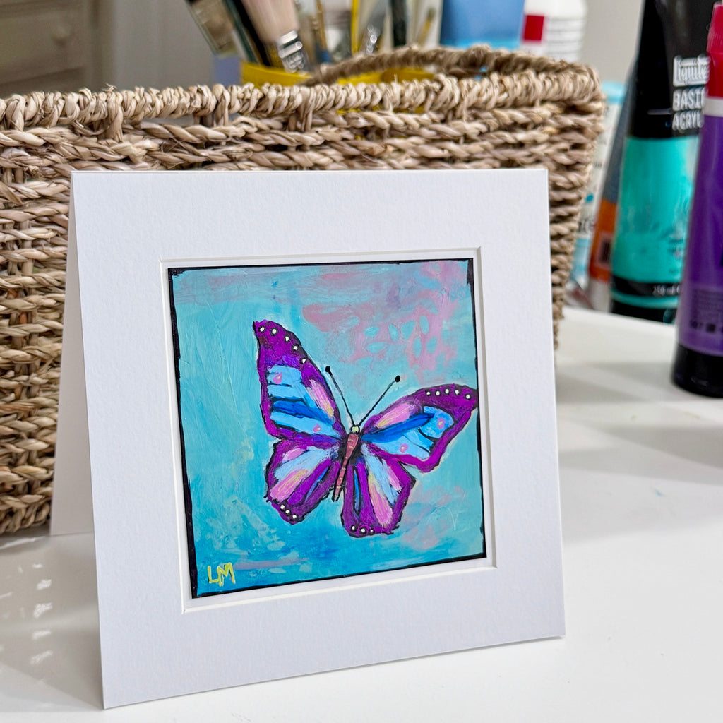 A butterfly painting in a white mat standing on a table with a basket, paints and brushes behind it
