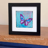 A butterfly painting in a black frame with a white mat standing on a table with text below it 'frame shown for display only, not included'