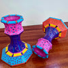 Two colorful hand-painted candle holders with geometric patterns, displayed on a wooden table against a plain wall.