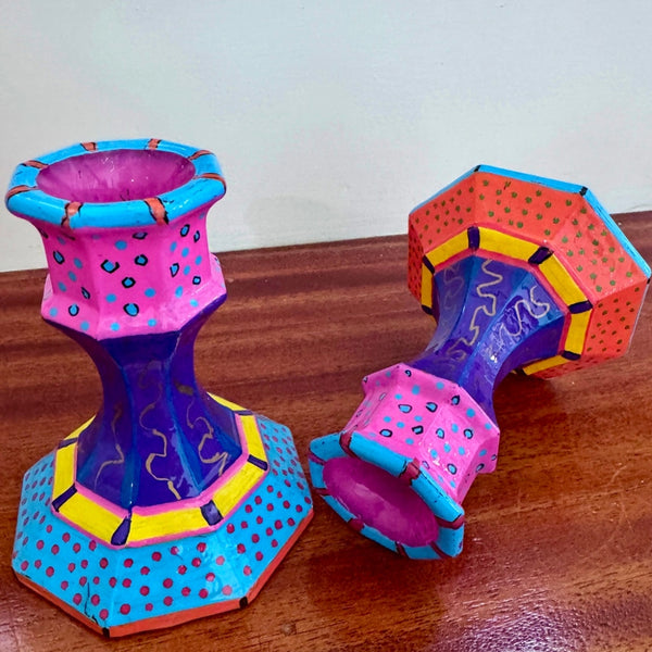 Two colorful hand-painted candle holders with geometric patterns, displayed on a wooden table against a plain wall.