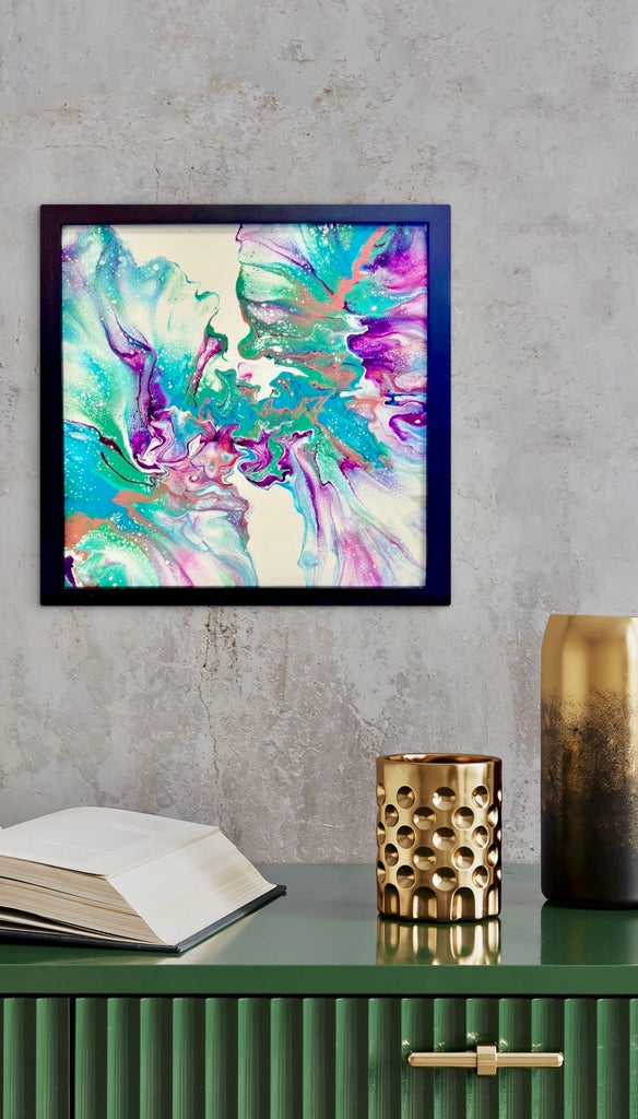 Carnival abstract painting in a black frame on a grey wall above a green cabinet with a book and gold decorative accents